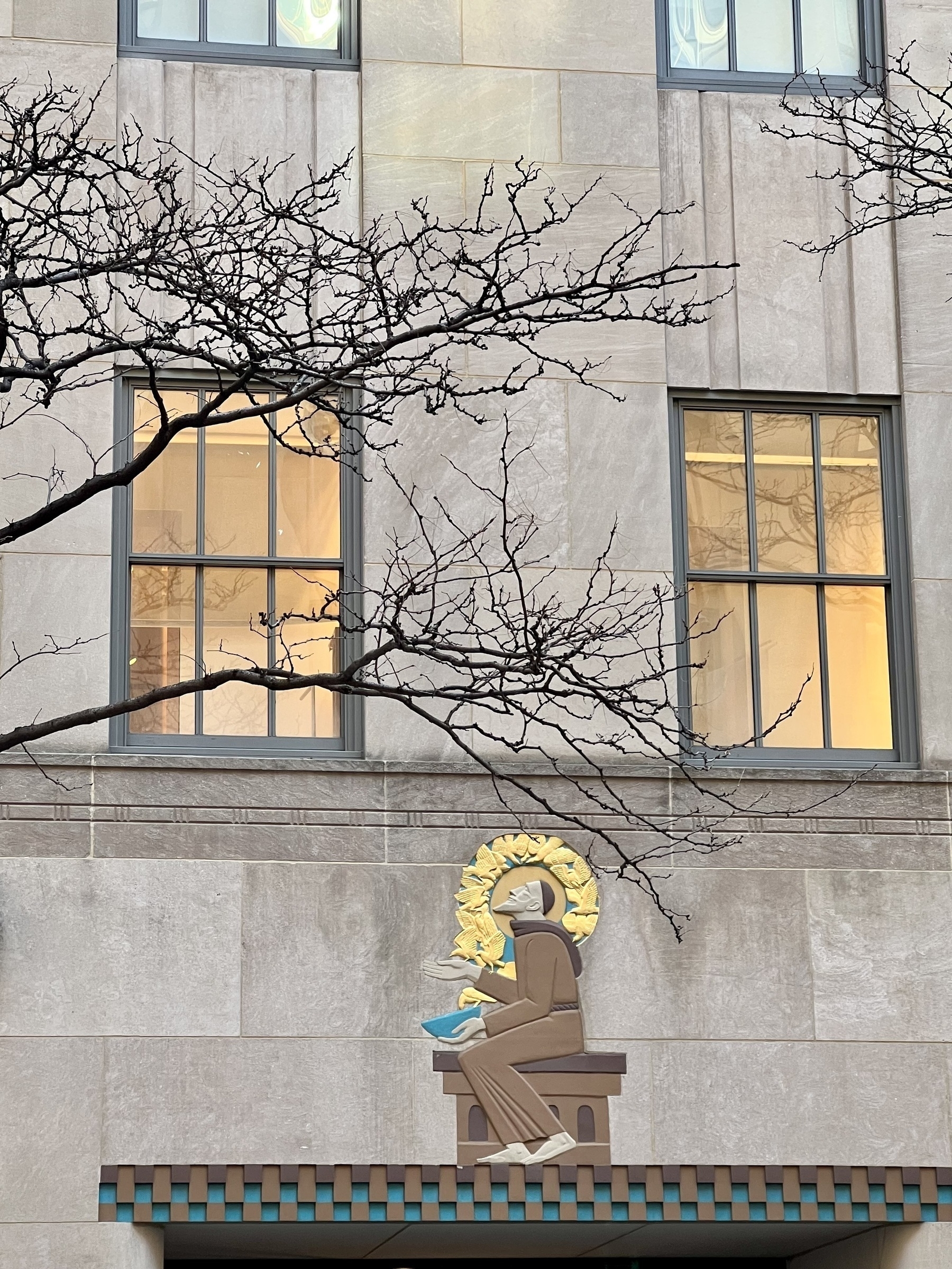 Bare tree branches frame a view of a building with warmly lit windows and an artistic depiction of Saint Francis seated and feeding birds on the wall outside.